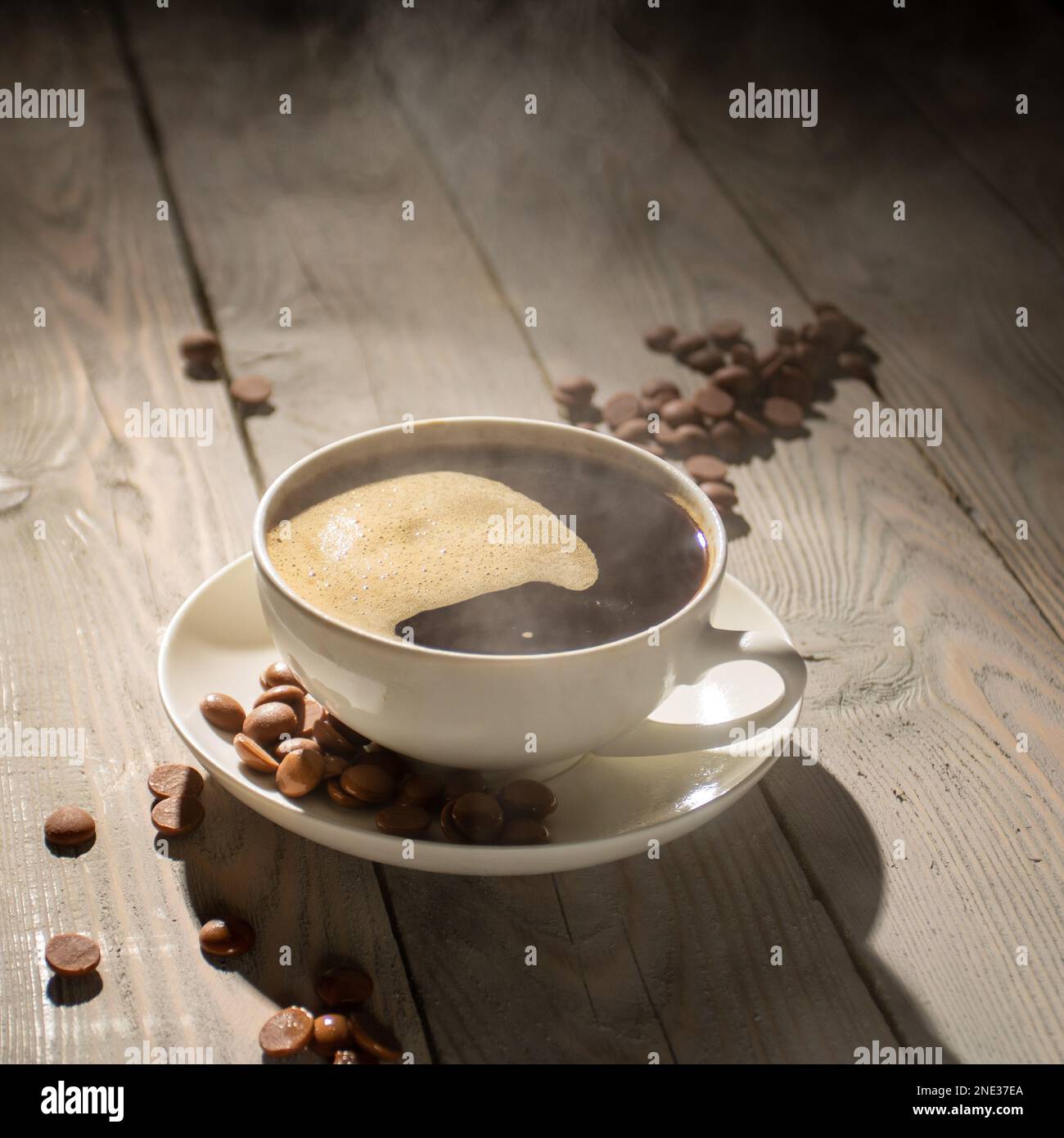 White cup of hot coffee with steam on a wooden table backlit. Stock Photo