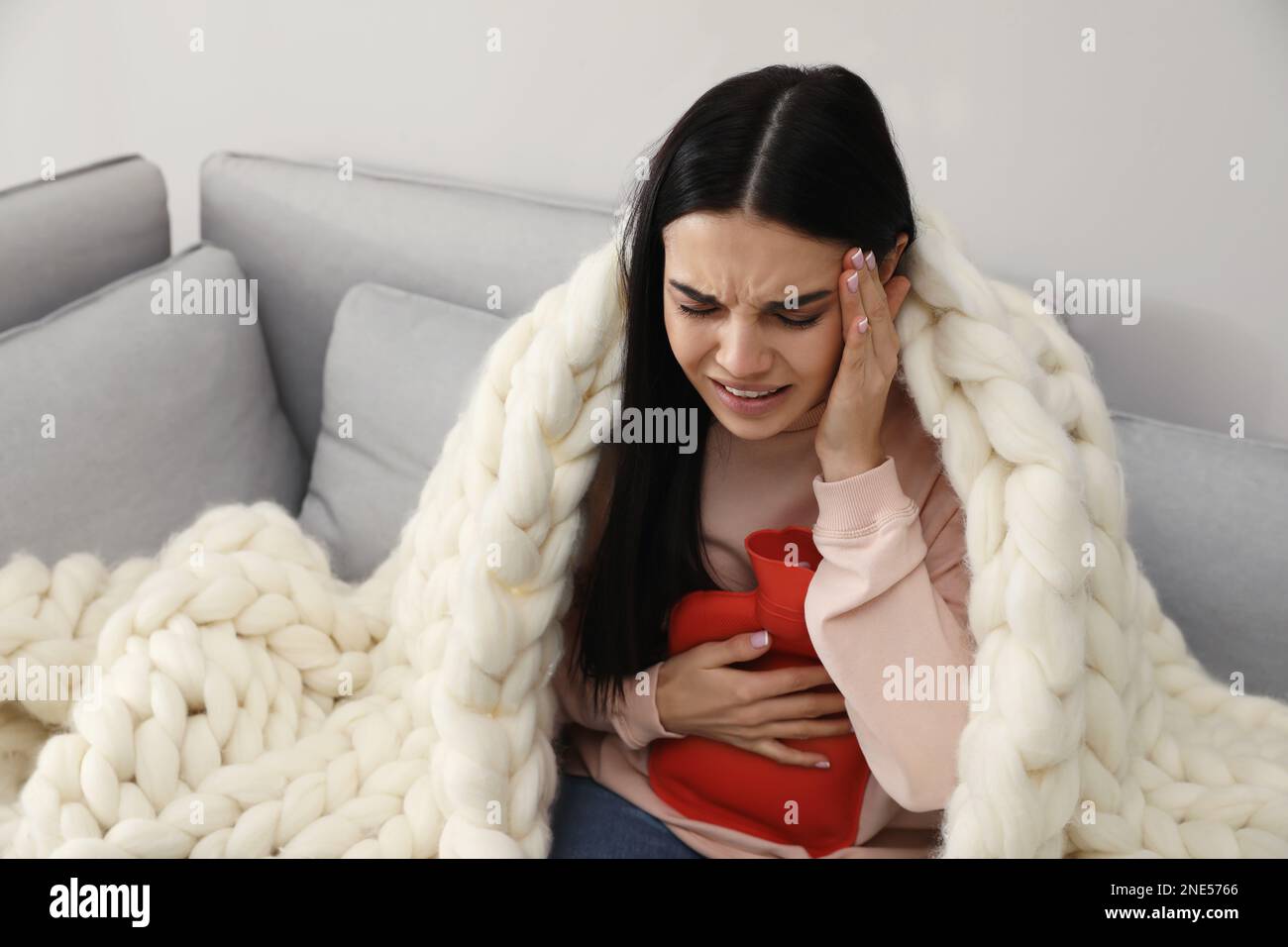 Woman using hot water bottle to relieve abdominal pain on sofa at home