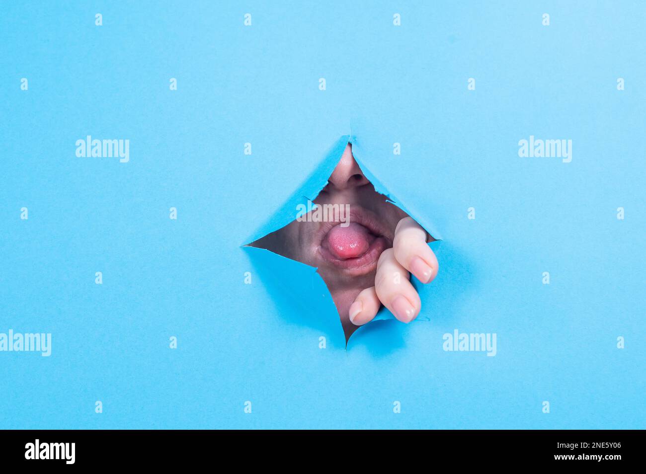 Woman sticking her tongue out of a hole on blue paper background.  Stock Photo