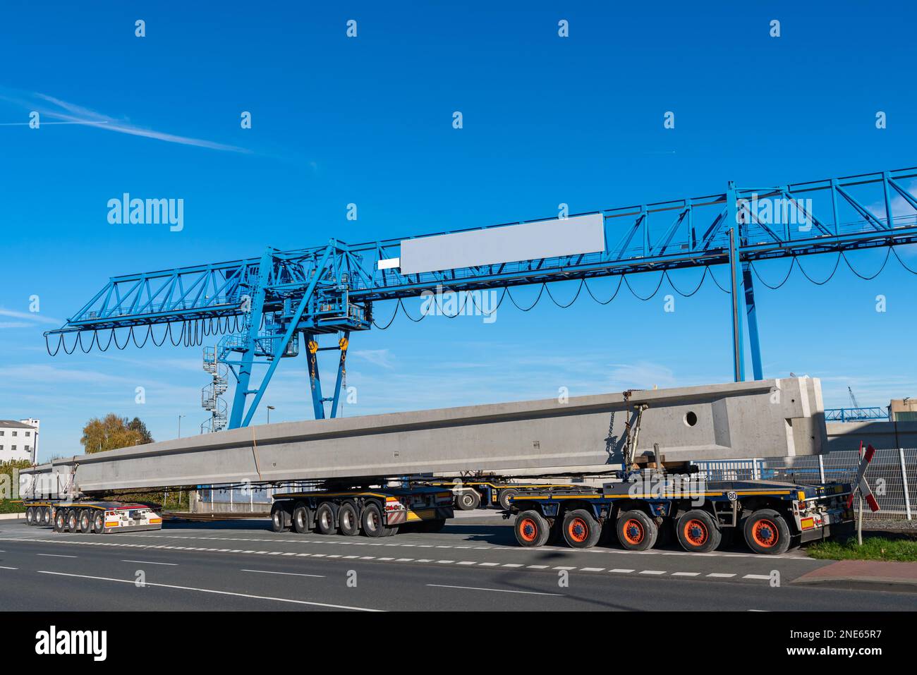 Overhead crane and trailer with long concrete beams against the blue sky. On the side of the road. Stock Photo