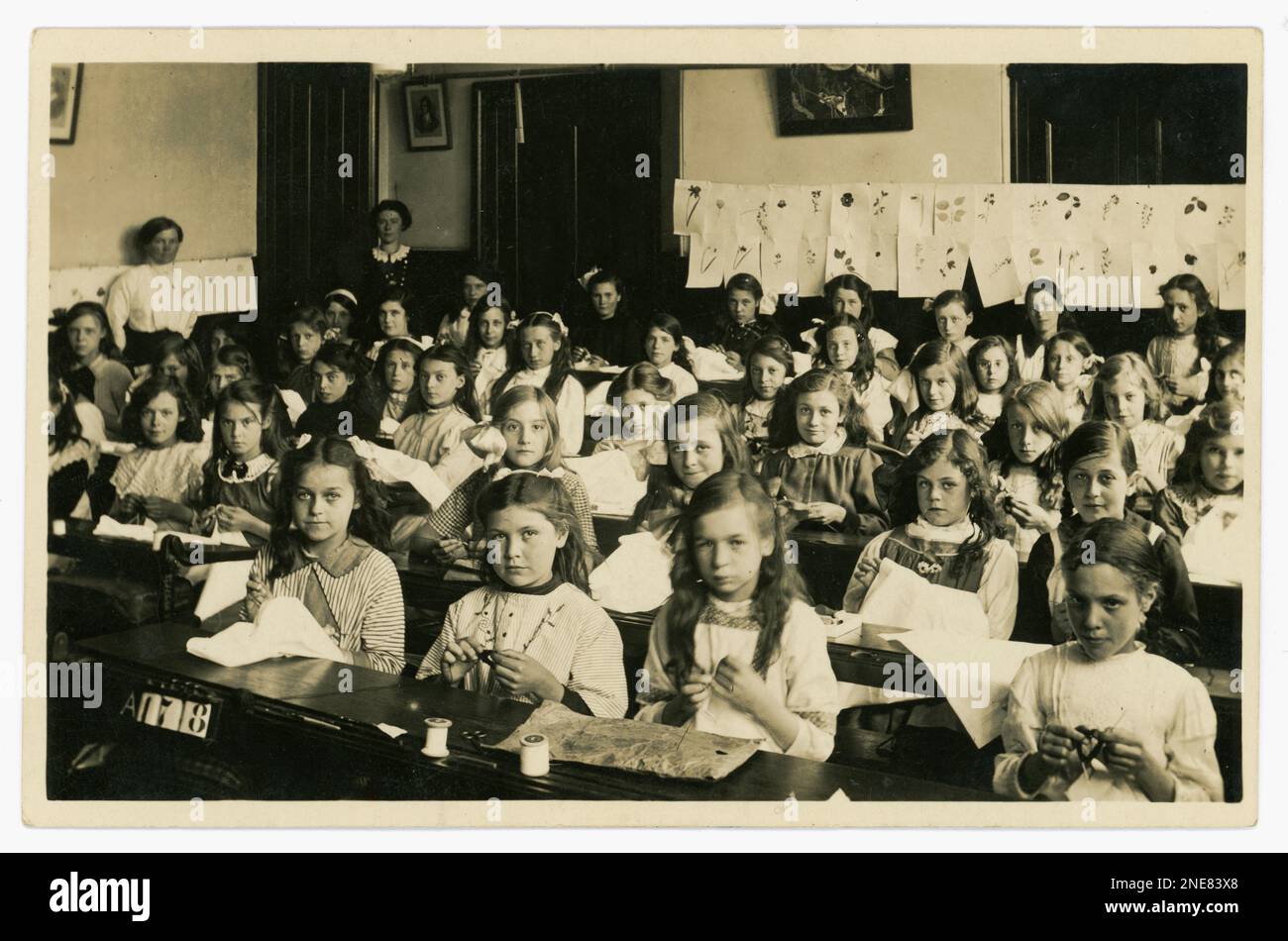 Original and clear WW1 era postcard of junior school girls in a sewing class, with samplers, possibly practising embroidery, some are knitting, lots of characters. No uniforms. Nature drawings on the wall. Two female teachers look on.  From the studio of J.& G Taylor, Green Lane, N. London, U.K.  Circa 1913-1919. Stock Photo