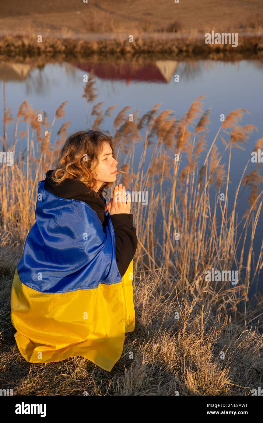 sad young patriot woman, wrapped in a Ukrainian flag, sits in despair on the shore of a lake and prays for peace for Ukraine. Russian aggression. suff Stock Photo