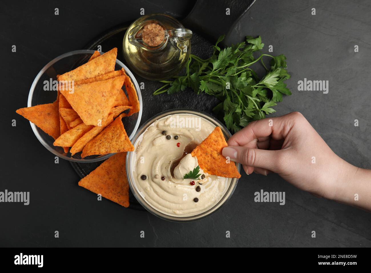 Woman dipping nacho chip into hummus at black table, top view Stock