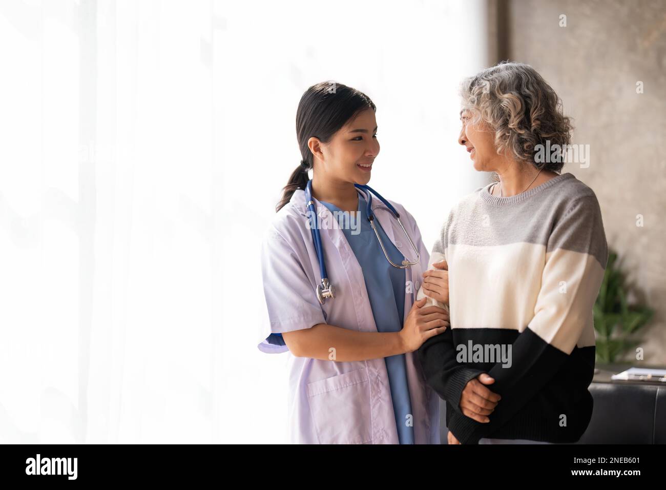 Young caregiver helping senior woman walking. Nurse assisting her old woman patient at nursing home. Senior woman with walking stick being helped by Stock Photo