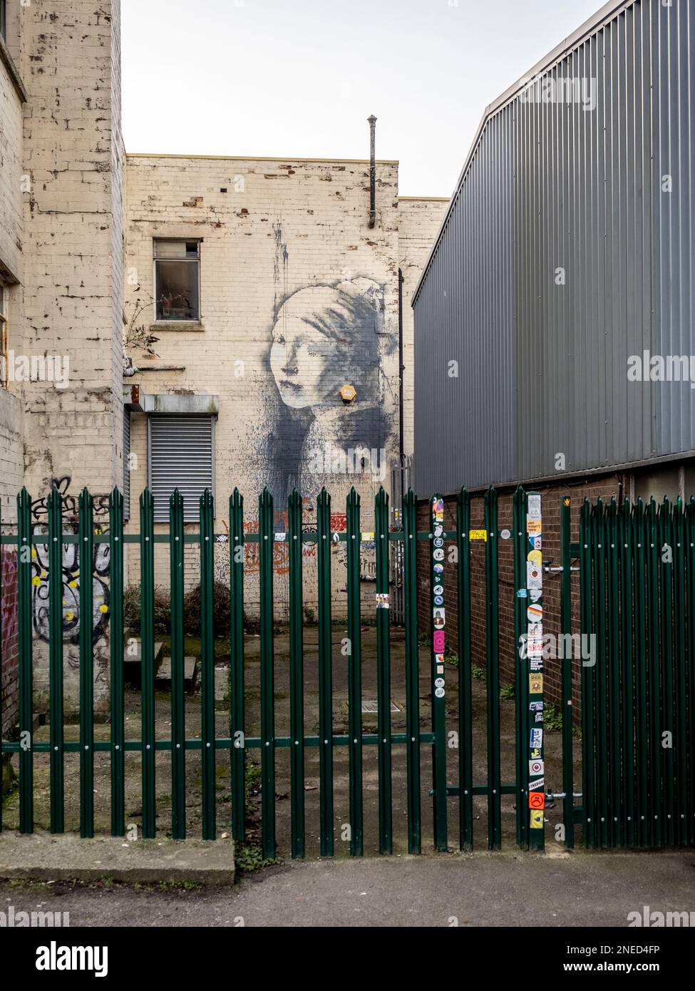 Banksy alleyway graffiti mural, The Girl with the Pierced Eardrum (inspired by Vermeer's painting) protected by a metal security fence. Bristol. UK Stock Photo