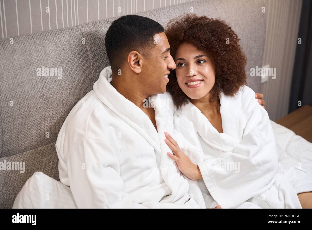 Smiling curly woman and her boyfriend are sitting embracing on bed ...