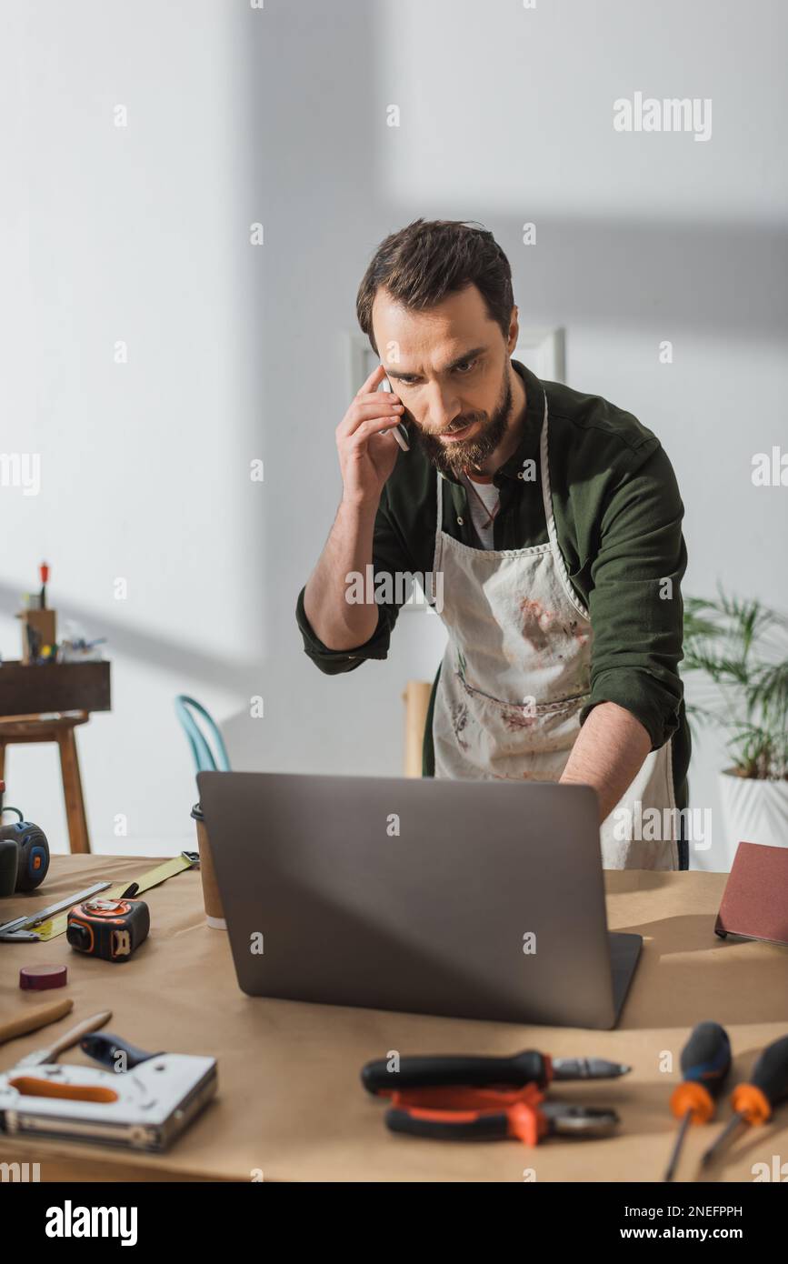 Bearded carpenter talking on smartphone and using laptop in workshop,stock image Stock Photo