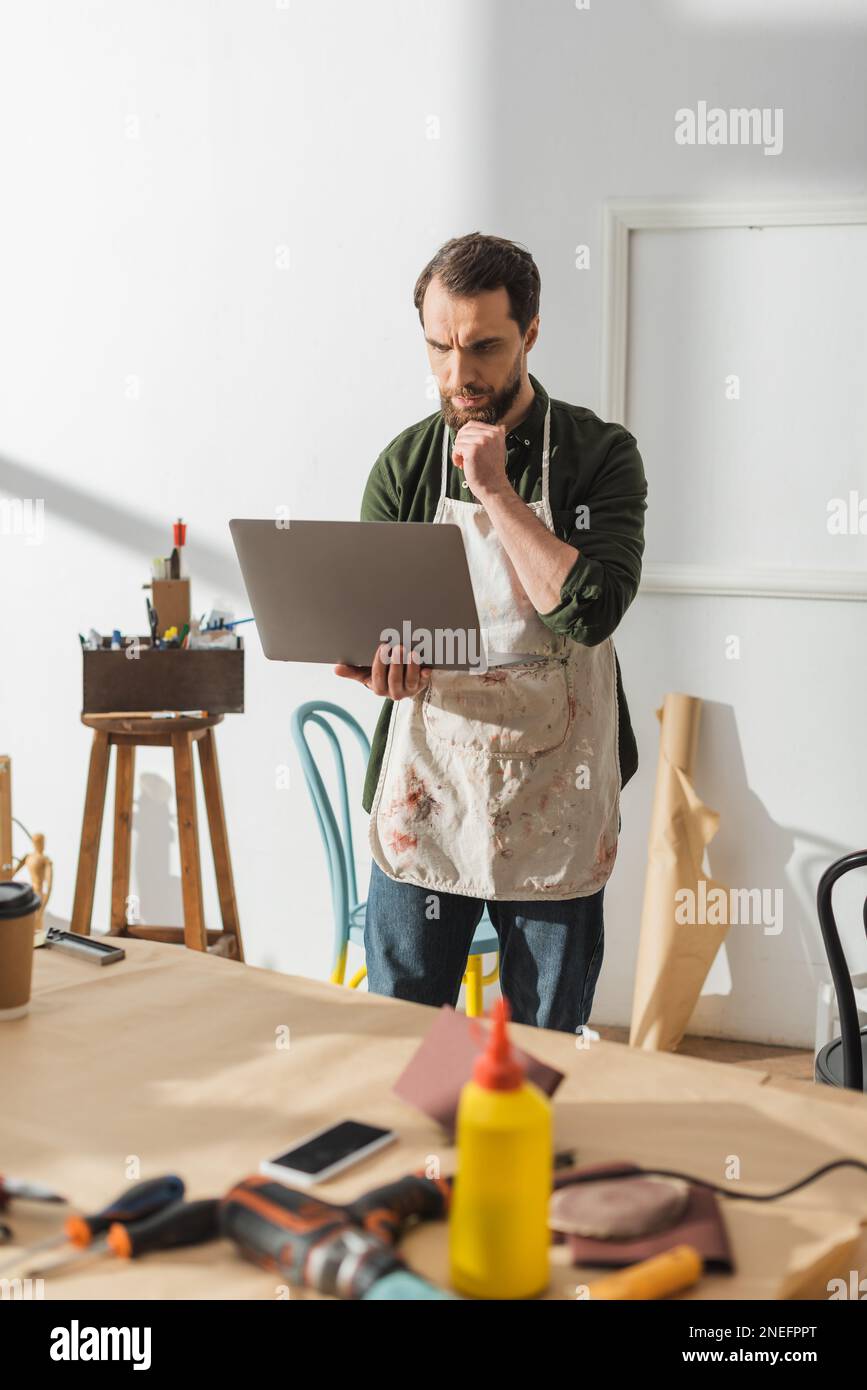 Pensive carpenter in apron using laptop in workshop,stock image Stock Photo