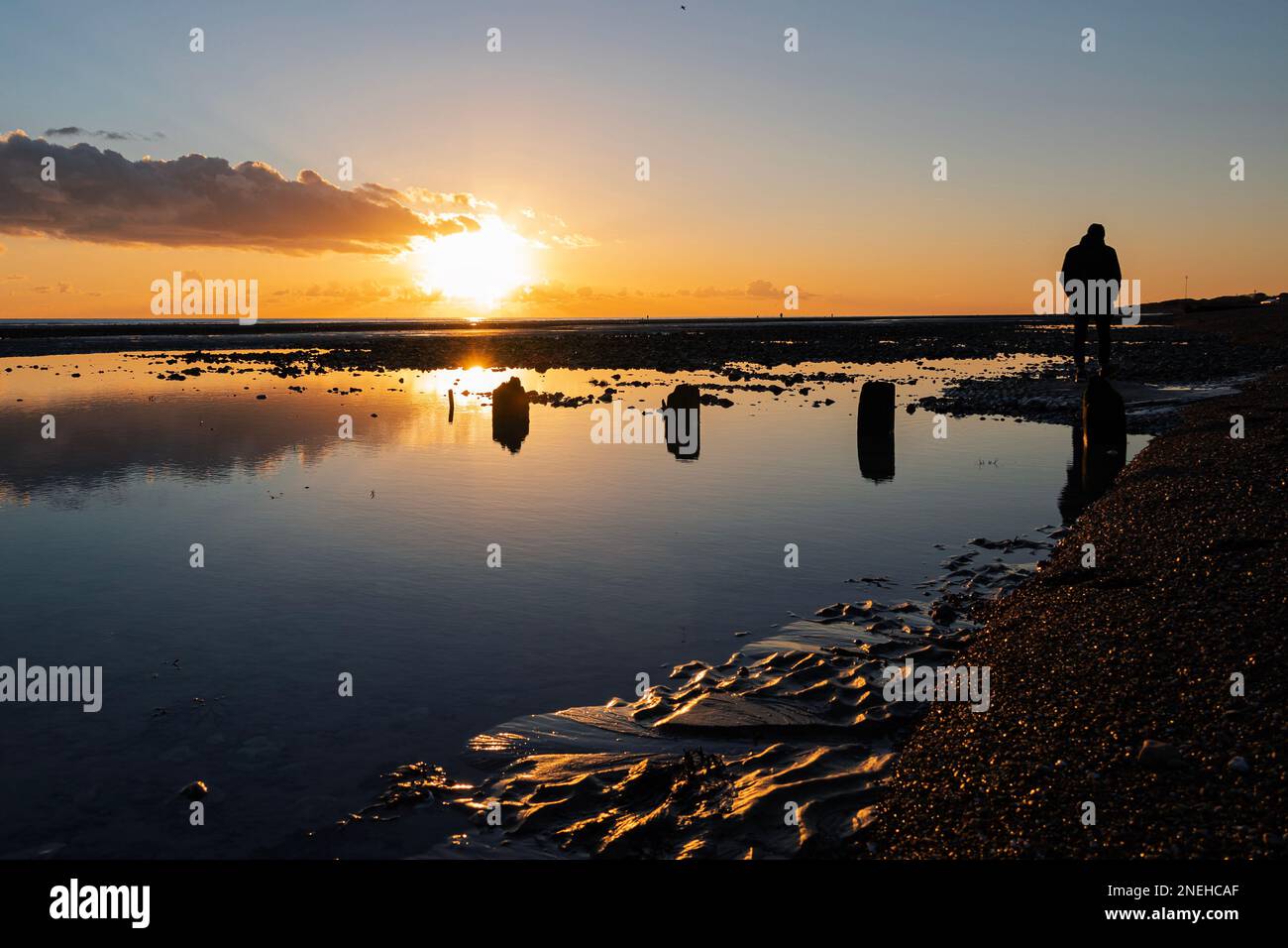Orange sky over Worthing, West Sussex in Winter with a man in silhouette walking along the beach Stock Photo