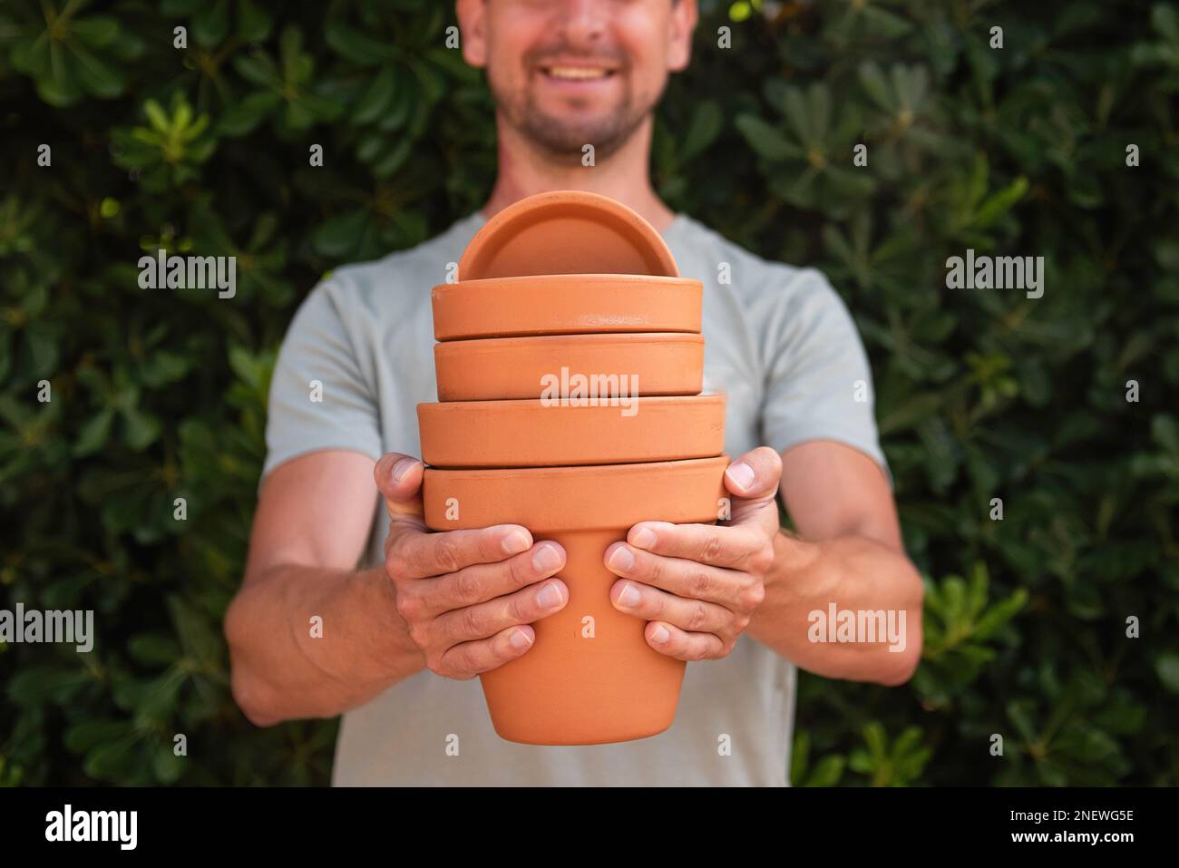 Close-up portrait man gardener in green cap holds terracotta clay pots in hands against the backdrop of an evergreen fence. Gardening, small business, Stock Photo