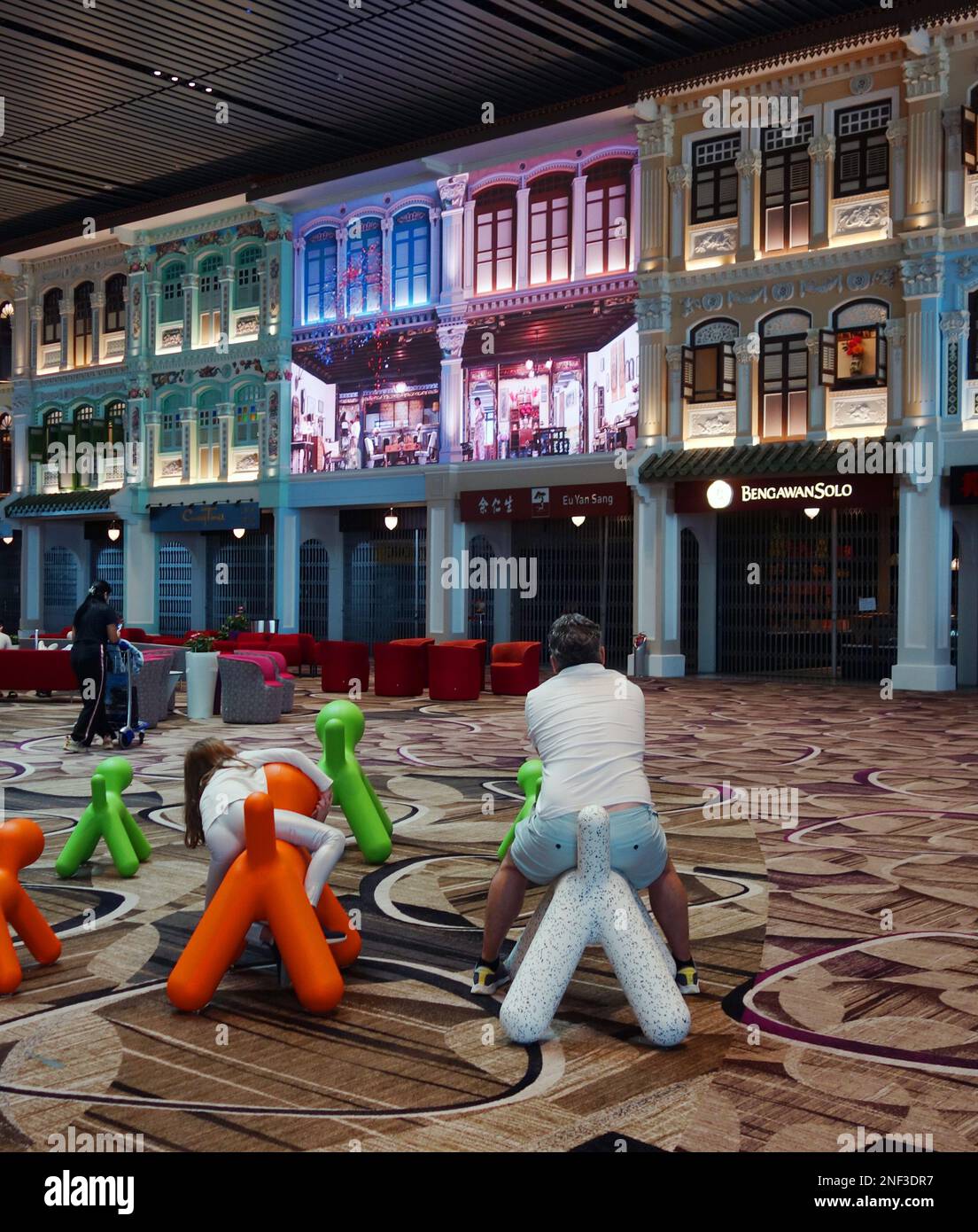 Family sitting on dog-shaped chairs watching projection of life in traditional town houses, Singapore airport, No MR or PR Stock Photo
