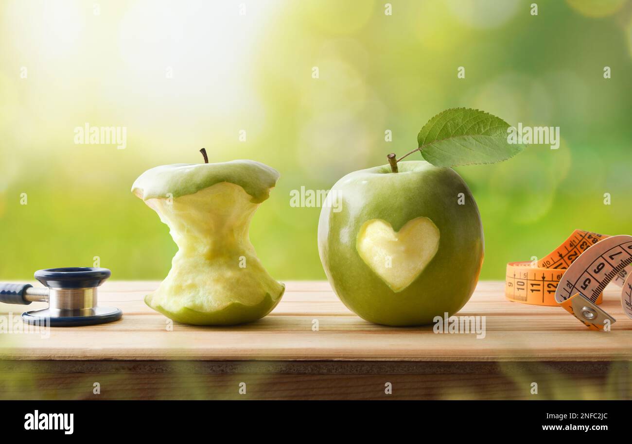 Healthy life and health check concept with eaten apple and whole apple with a bitten heart, stethoscope and tape measure on wooden table with nature b Stock Photo