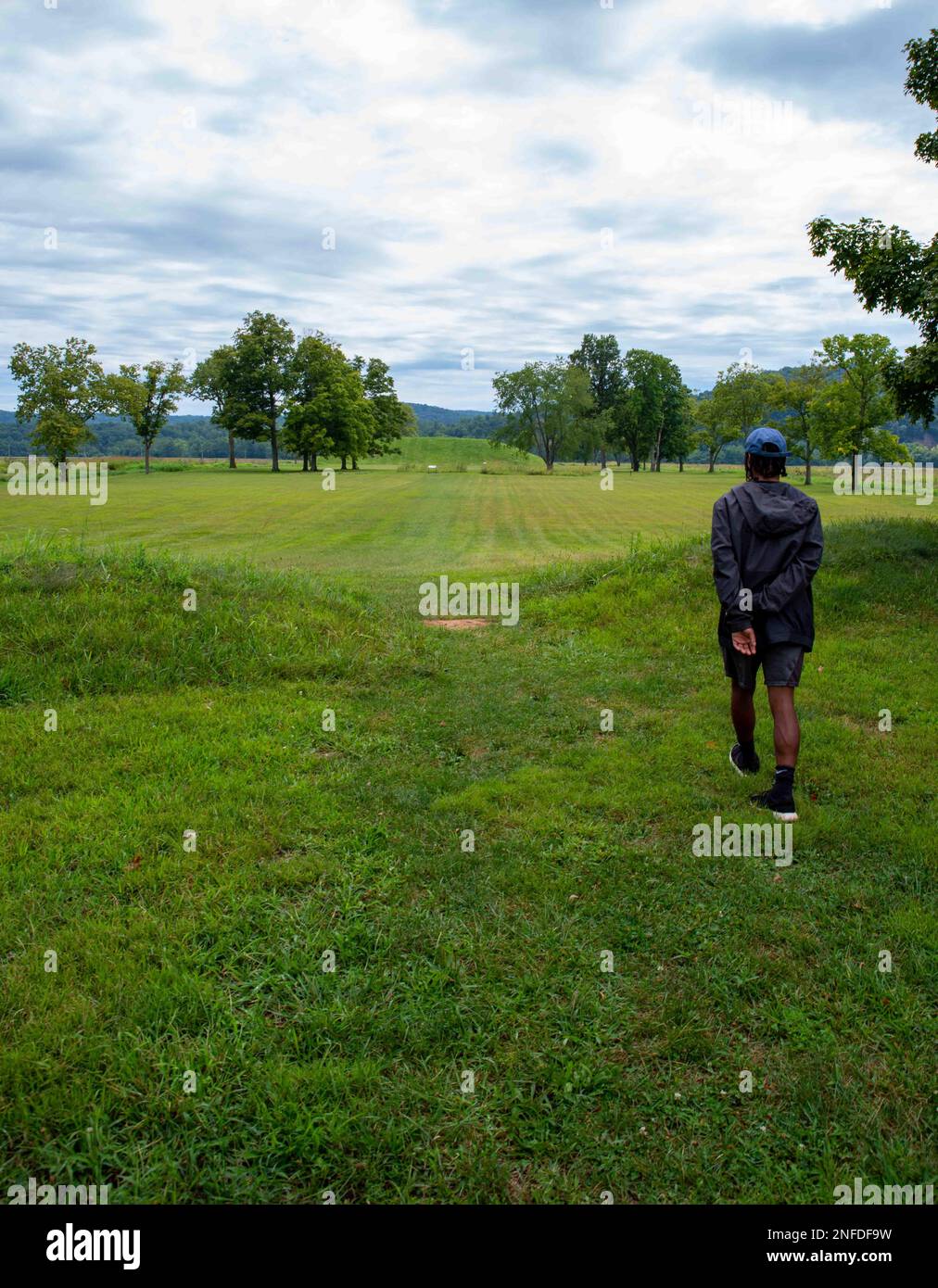 A young man walks toward the Native American Hopewell Culture prehistoric Seip Earthworks burial mound in Ohio. Ancient large long mound. Grass is nea Stock Photo