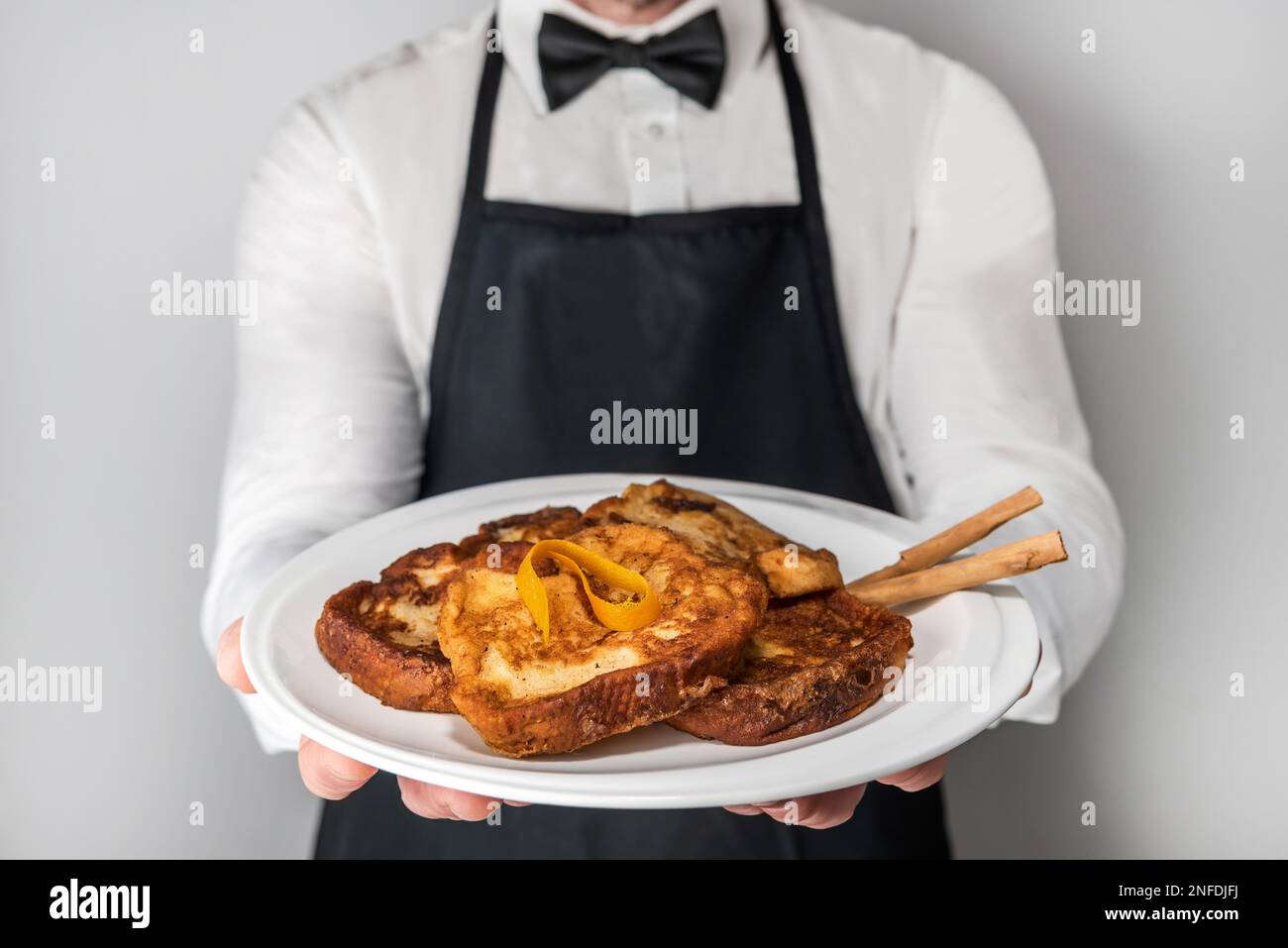 A chef waiter in apron and bow tie showing a plate of gourmet torrijas. A typical Spanish sweet made with bread, milk, eggs, sugar and cinnamon that i Stock Photo