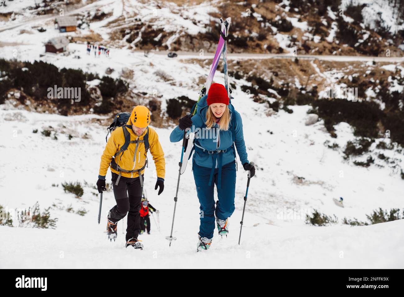 Couple of mountaineers climbing up a snowy mountain, woman carrying skis on her back  Stock Photo