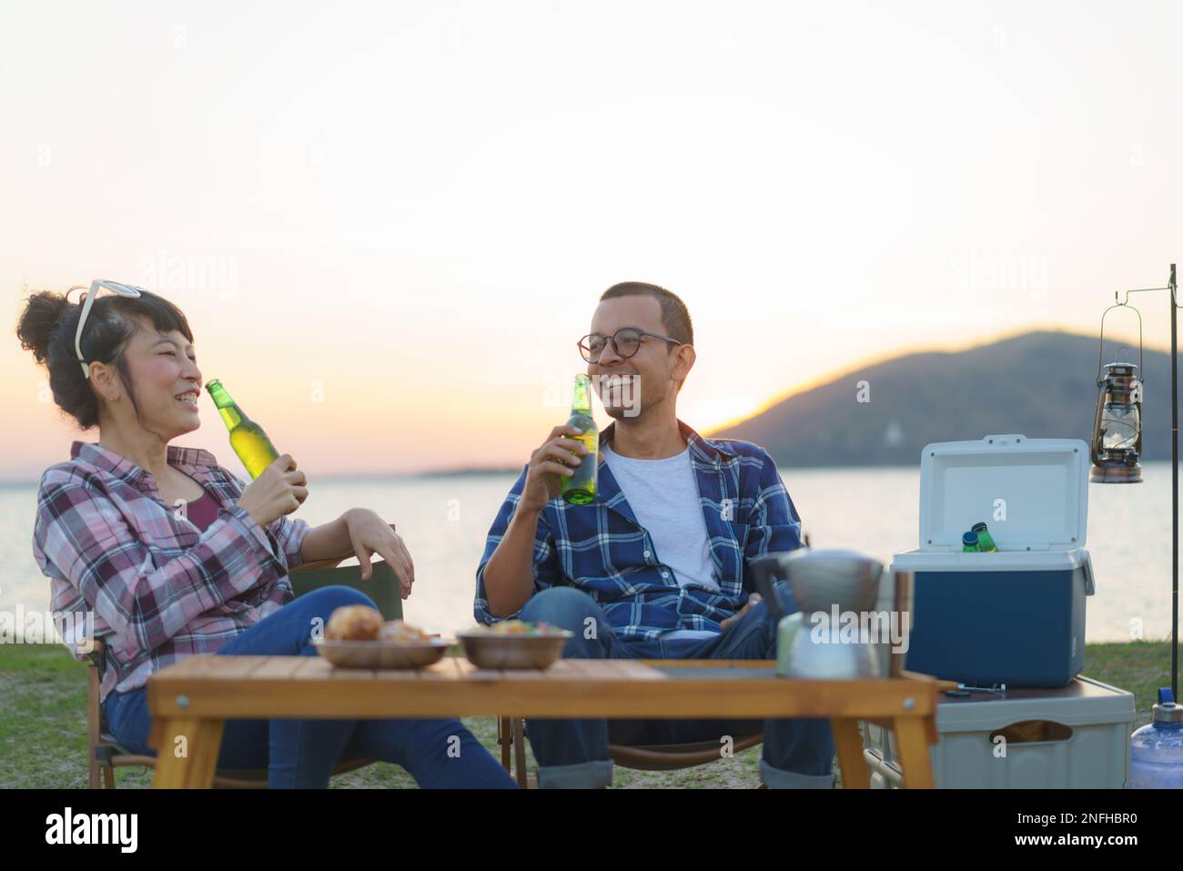 Asian couple drinking beer from bottle in their camping area with lake in the background during sunset. Stock Photo