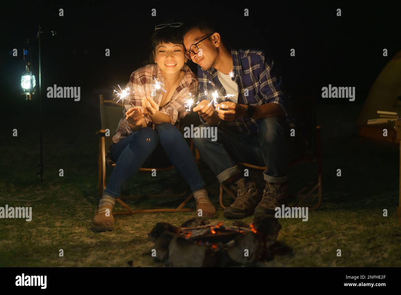 Asian couple is lighting sparkler fire at a campfire where they set up a tent to camp by the lake at night. Stock Photo