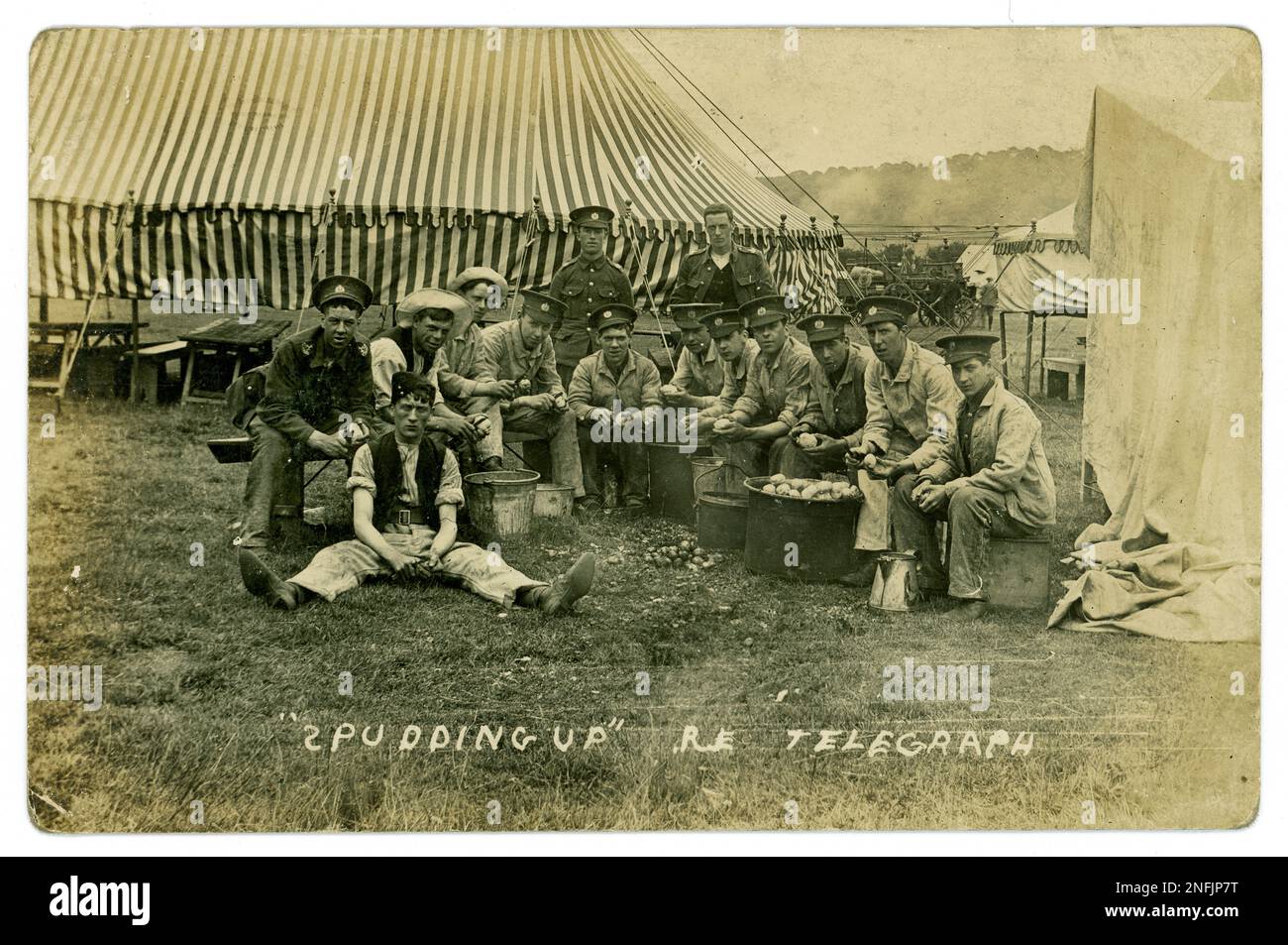 WW1 postcard of group of  Royal Engineers in Telegraph Corps, peeling potatoes next to the camp dining tent, entitled 'Pudding Up R.E. Telegraph'. Three of the men have the Lincolnshire Territorial corps visible and the rest are probably the same but their uniform is covered up with a work jacket.. A supply truck is in the background looked over by civilians. Circa 1915, possibly a training camp in Lincolnshire, U.K. Stock Photo