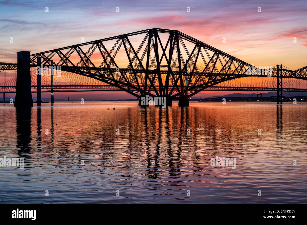 Colourful reflections of the Forth Rail Bridge at dusk, with Forth Road Bridge and Queensferry Crossing beyond, from South Queensferry, Scotland, UK Stock Photo