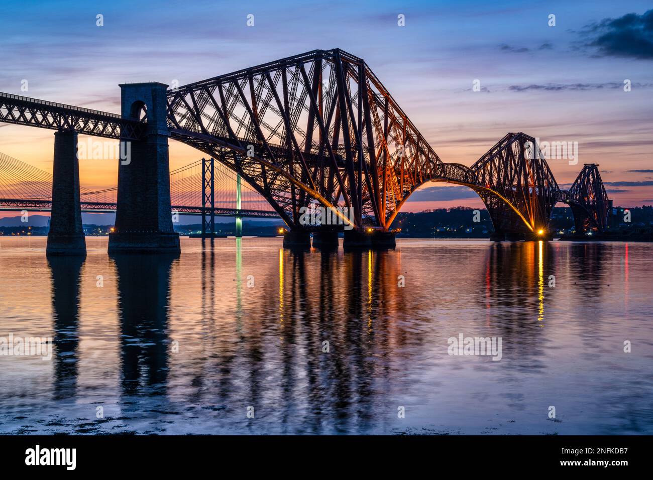 Illuminated Forth Rail Bridge at dusk, with Forth Road Bridge and Queensferry Crossing beyond, from South Queensferry, Scotland, UK Stock Photo