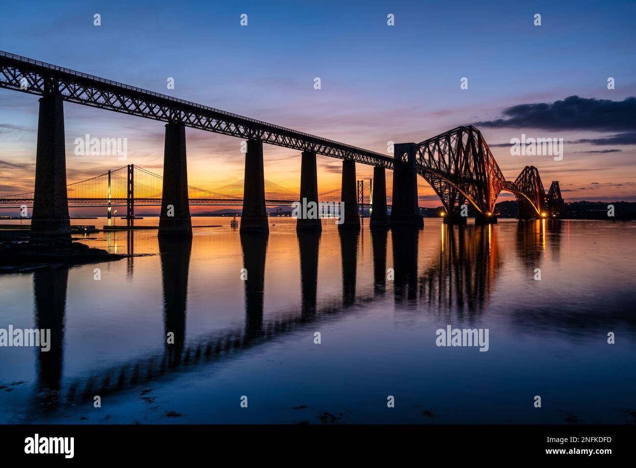 Illuminated Forth Rail Bridge at dusk, with Forth Road Bridge and Queensferry Crossing beyond, from South Queensferry, Scotland, UK Stock Photo