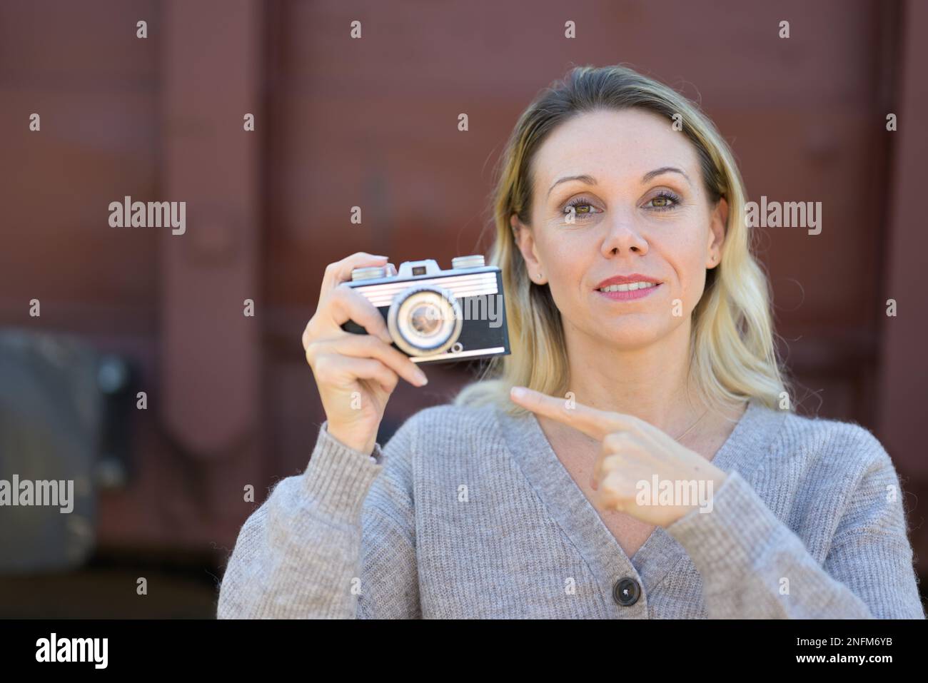 Pretty middle aged woman holding an old camera and pointing a finger at the camera wearing a brown sweater in front of an old train wagon Stock Photo
