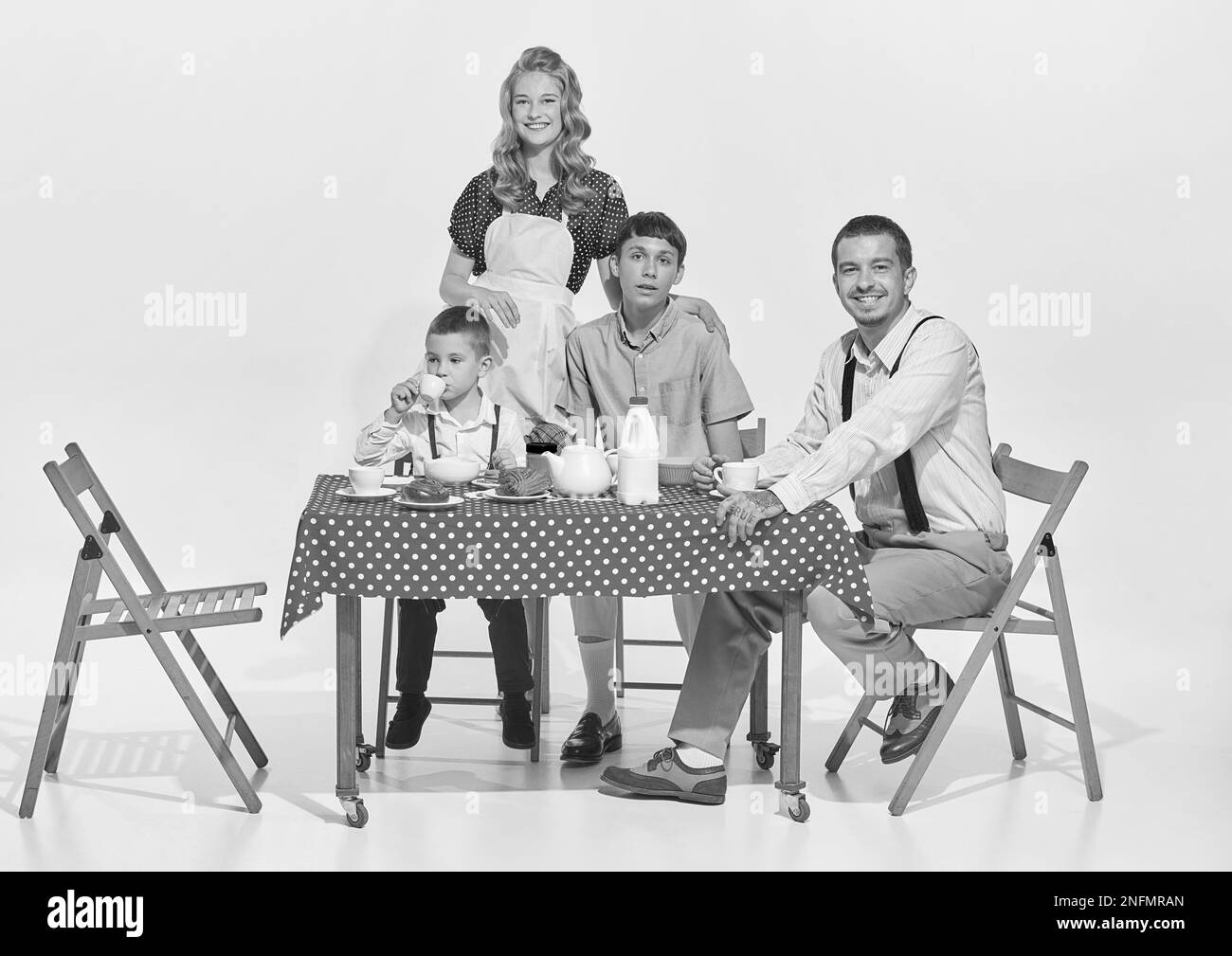 Portrait of lovely young family, woman, man and two boys sitting at the table and having breakfast isolated over grey background. Morning talk Stock Photo