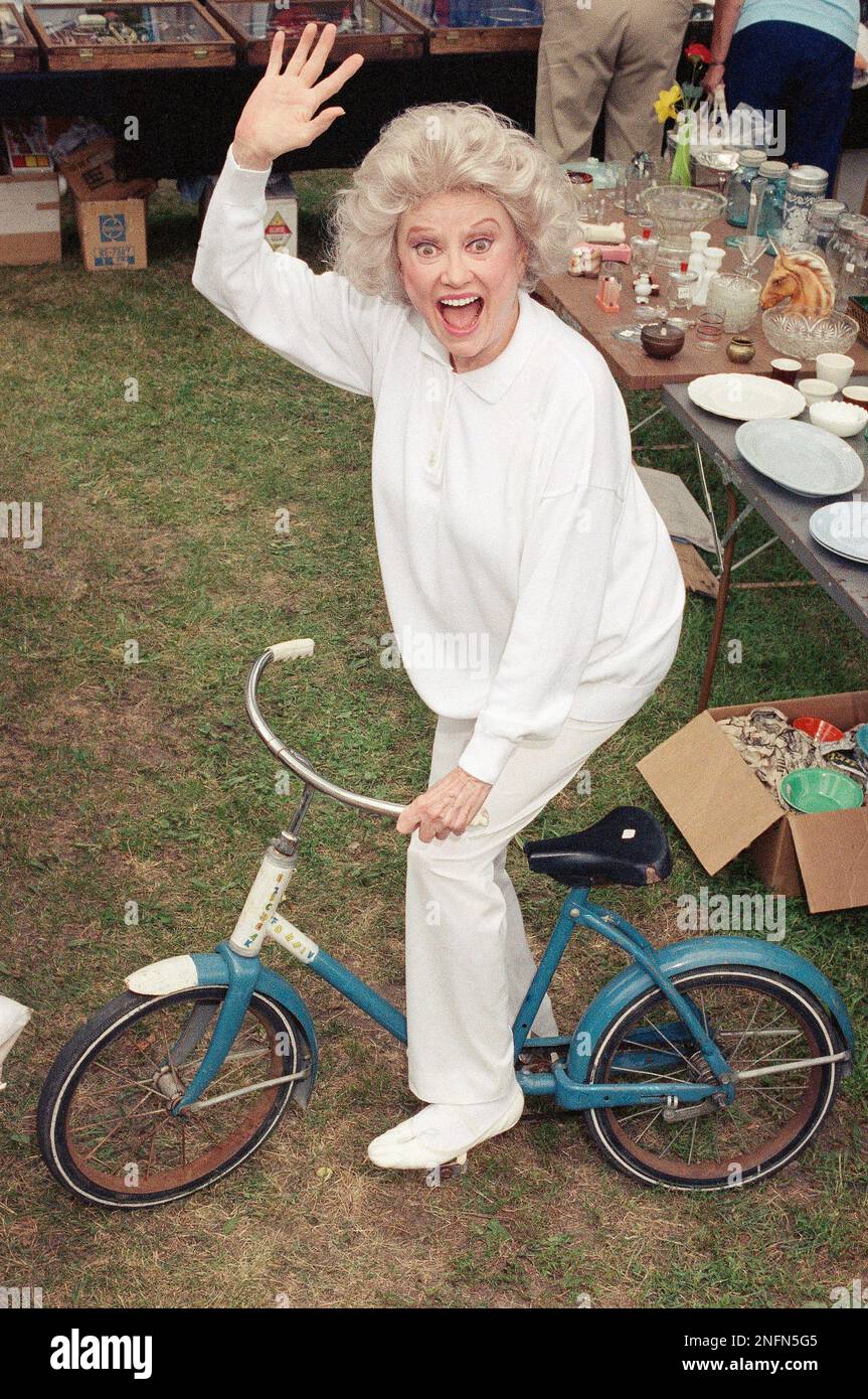 Entertainer Phyllis Diller clowns around on a bicycle at a garage sale
