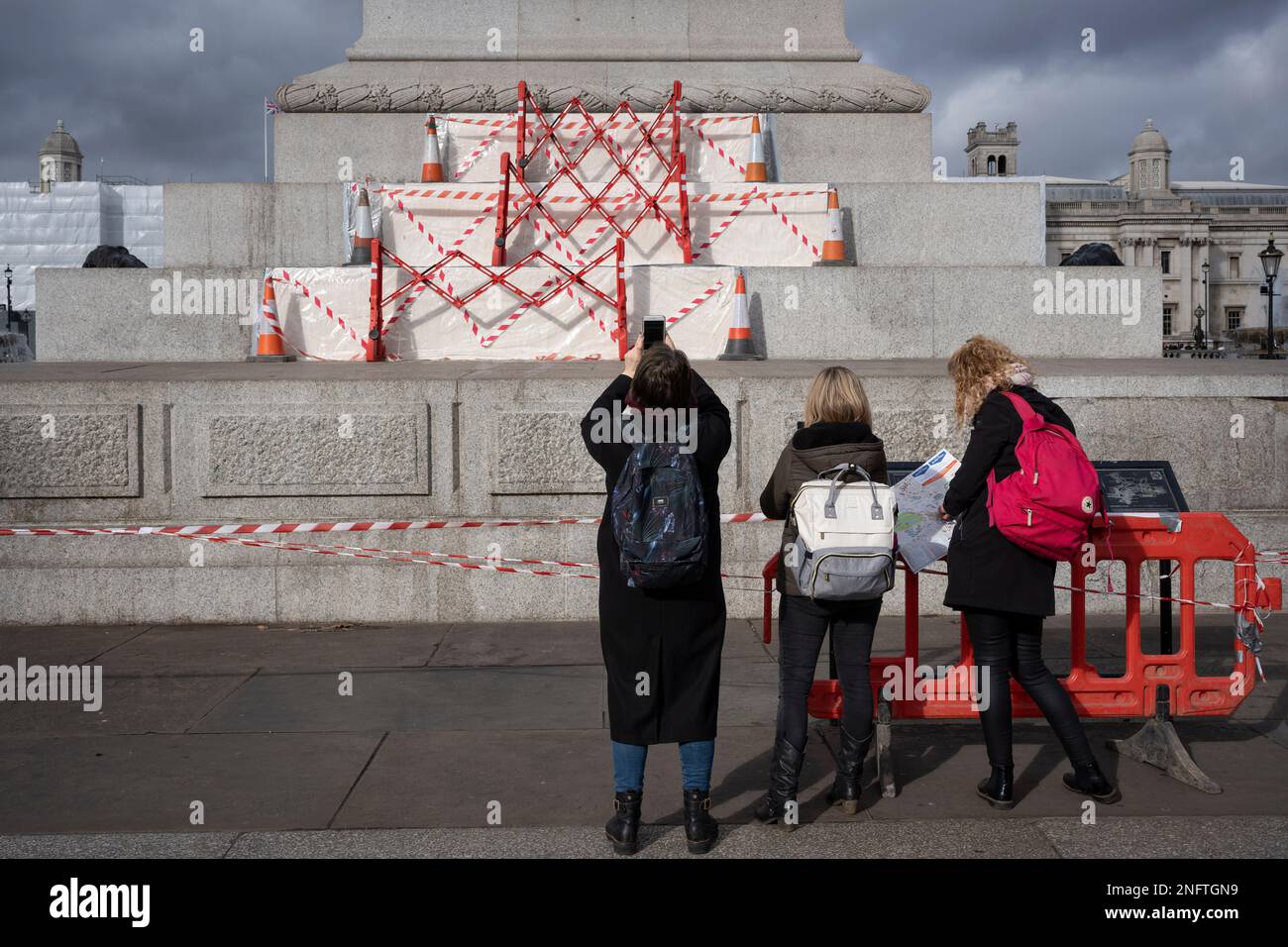 Members of the public seemingly admire hazard tape that covers repairs to granite stonework of the plinth to Nelson's Column in Trafalgar Square, on 17th February 2023, in London, England. Stock Photo