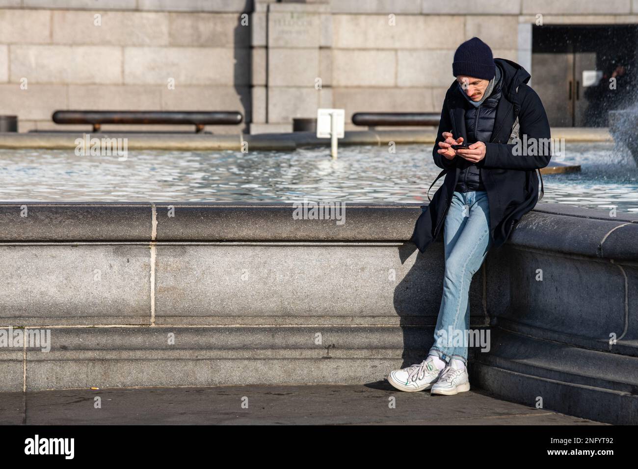 Man browsing his mobile phone by fountain at Trafalgar Square on a sunny winter day in London, England Stock Photo