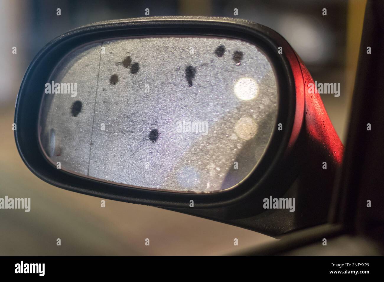Lef side mirror on car at night reflects a city lights and  water drops. Close up view of bright blurred background. Stock Photo