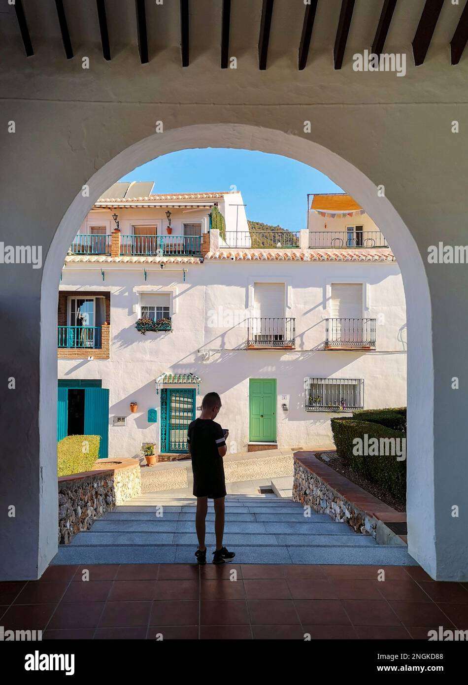 A young boy checking his phone below an arch in the white village of Frigiliana, Malaga Province, Andalucia, Spain. Stock Photo