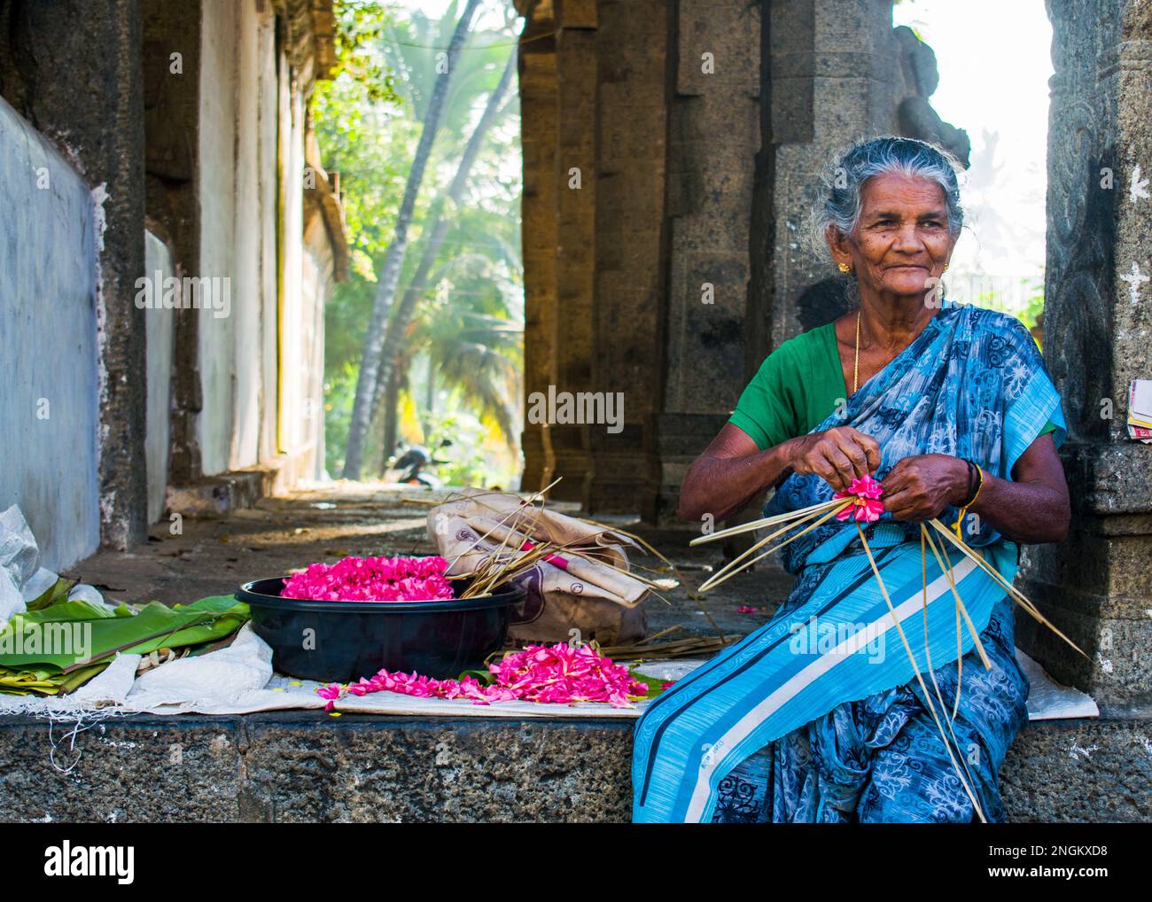 An old but happy Indian women weaving a flower garland in front of a