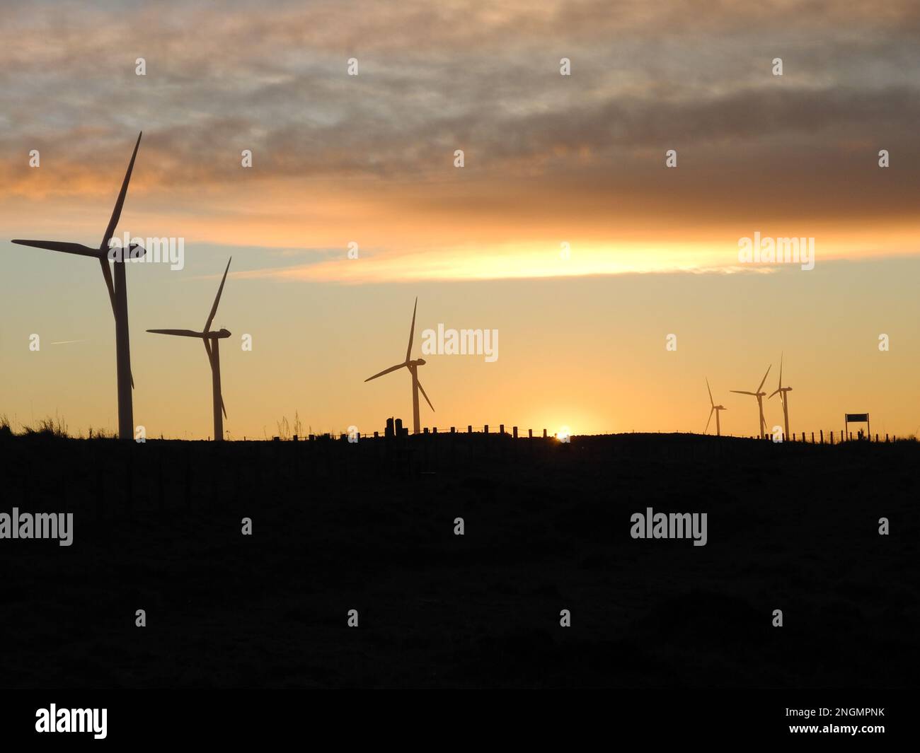 Wind turbines in silhouette against the warm glow of the setting sun on the horizon and with a dark foreground Stock Photo