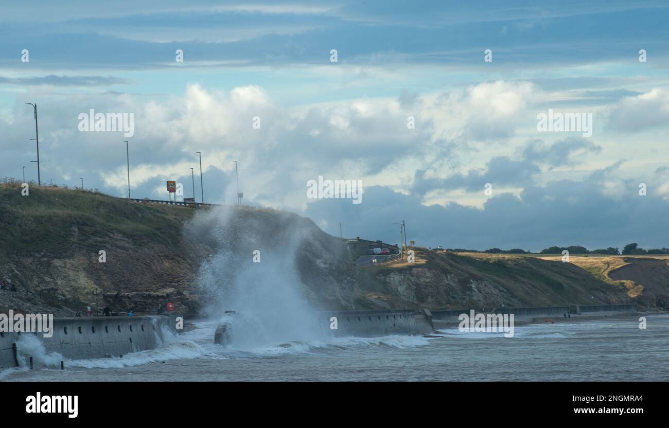 Heavy waves crash onto the promenade at Seaham throwing up columns of spray with cliffs and coastal road caught in the sunlight Stock Photo