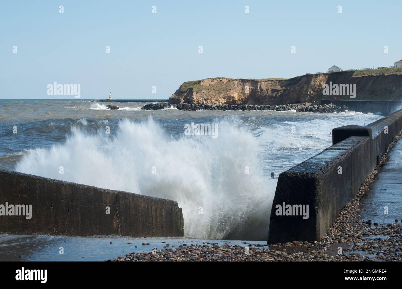 View along the promenade at Seaham including cliffs and distant lighthouse with waves crashing into the stonework throwing up plumes of spray Stock Photo