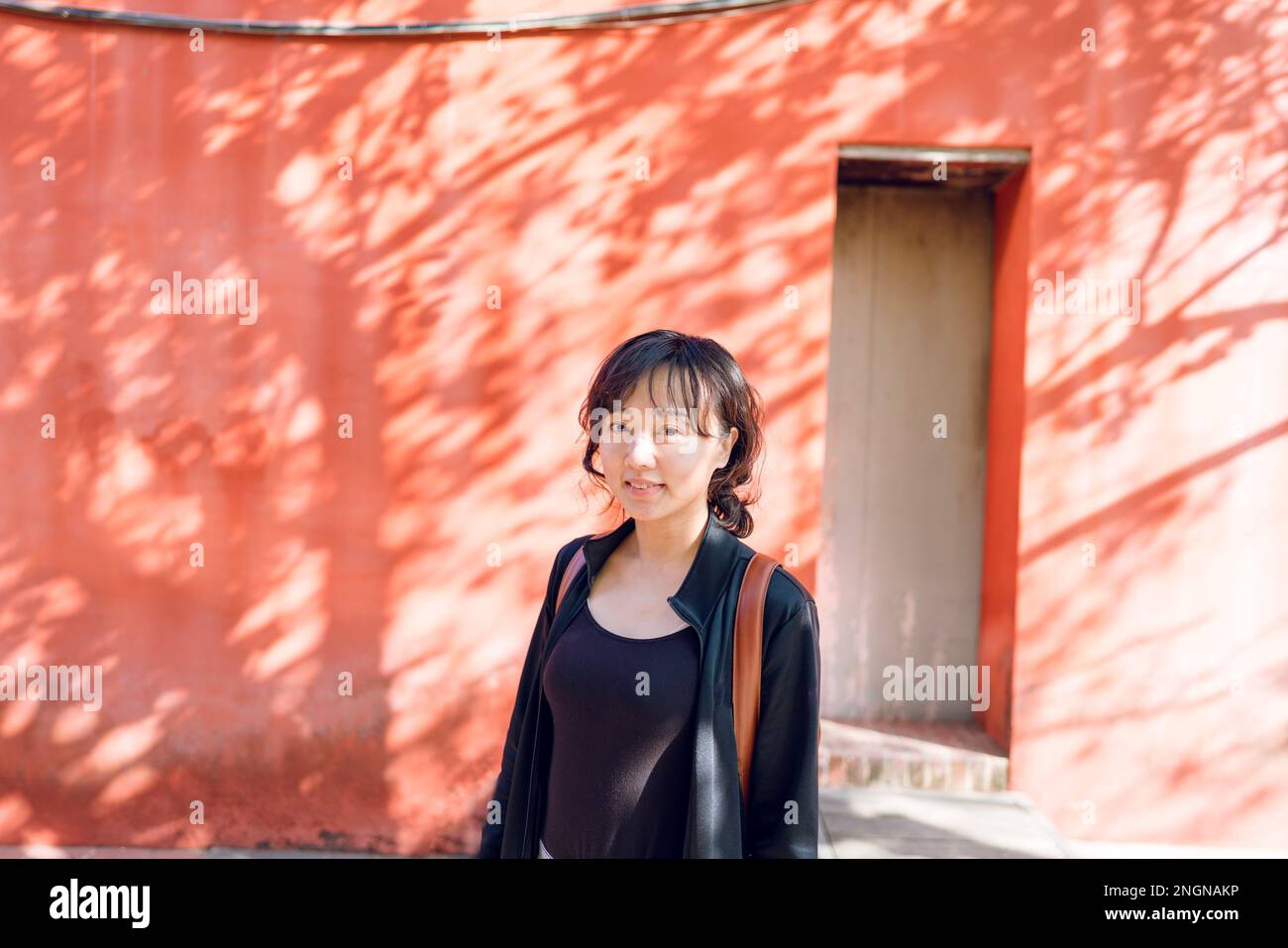 A woman standing in front of a red wall smiling at the camera. The trees shadows covered on the wall. Stock Photo