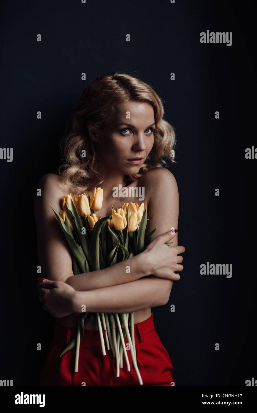Adult beauty woman half naked in formal evening red trousers without bra hugs bouquet of yellow tulips. Stylish blonde curly hair sensual nude model f Stock Photo
