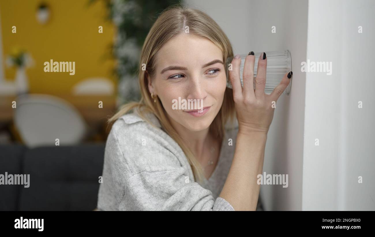 Young blonde woman listening through wall with glass at home Stock