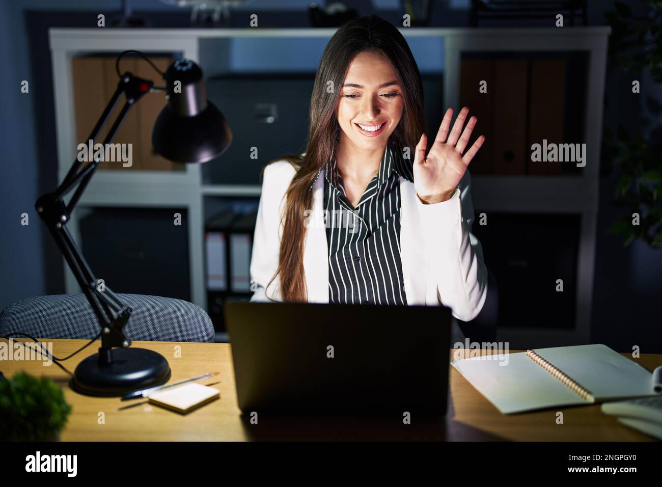 Young brunette woman working at the office at night with laptop waiving ...