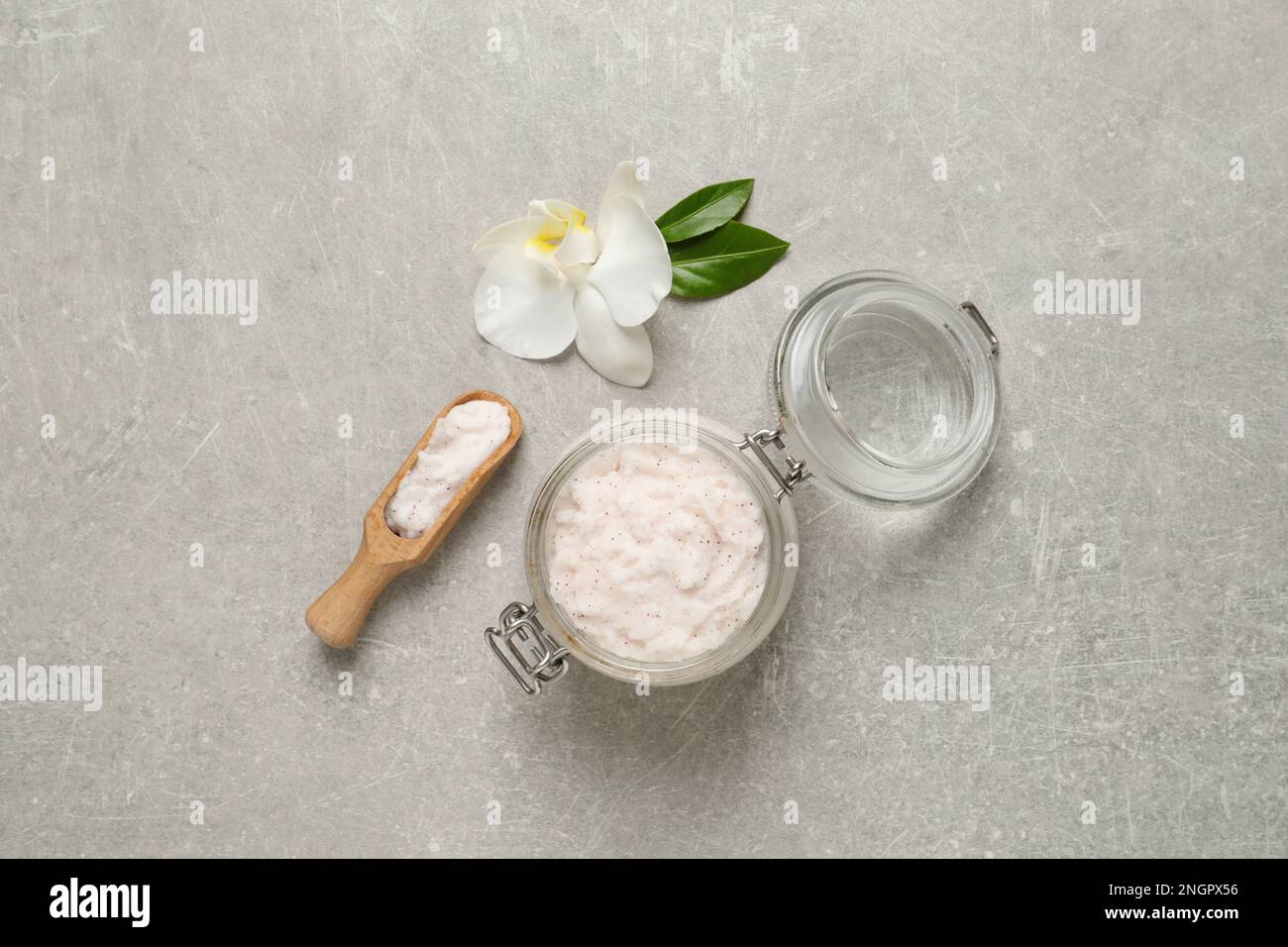Body scrub in glass jar and scoop near orchid flower on light grey