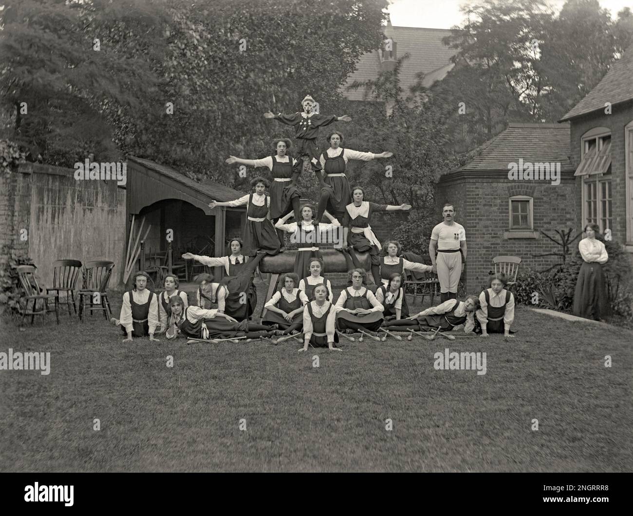 A gymnastic display outdoors with 18 schoolgirls, a young man and a male instructor forming a human pyramid posing on a ‘horse’, UK c.1900. Balance skills plus fitness and flexibility are called upon. Oddly, the young man in the top position wears a clown outfit. The male teacher is standing on the right. The other girls are dressed in long skirts and knickerbockers. In front of the girls are gymnastic clubs. The long exposure means there is some motion blur – a vintage 1800s/1900s, Victorian/Edwardian photograph. Stock Photo