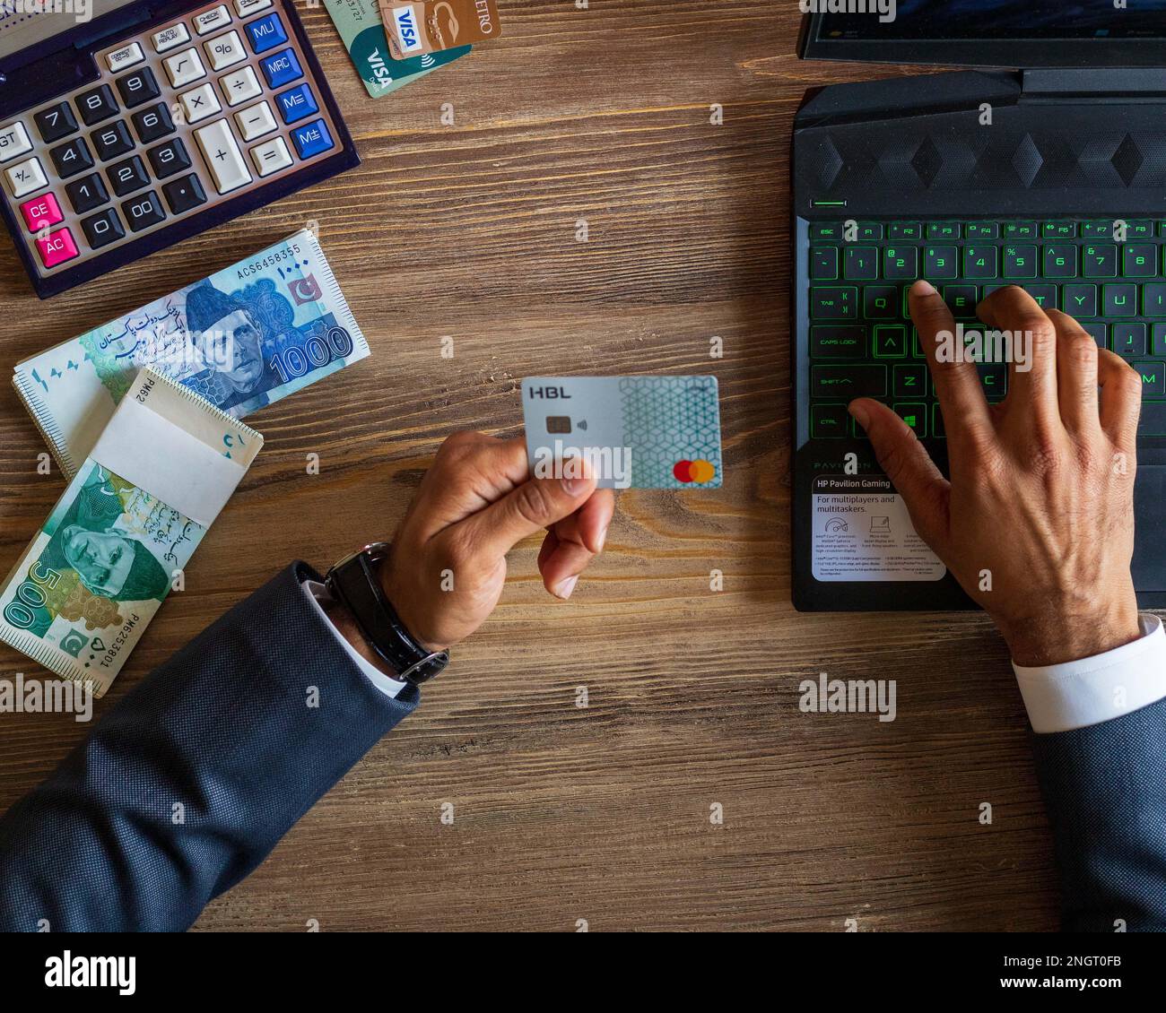 Business man using HBL bank debit card for online/ e-commerce transaction on his laptop, stack of Pakistani currency notes and calculator on table Stock Photo