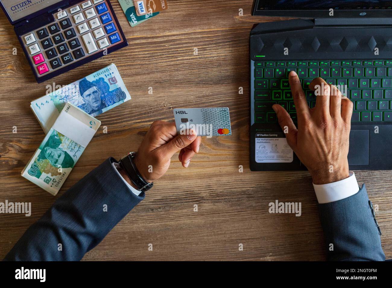 Business man using HBL bank debit card for online/ e-commerce transaction on his laptop, stack of Pakistani currency notes and calculator on table Stock Photo