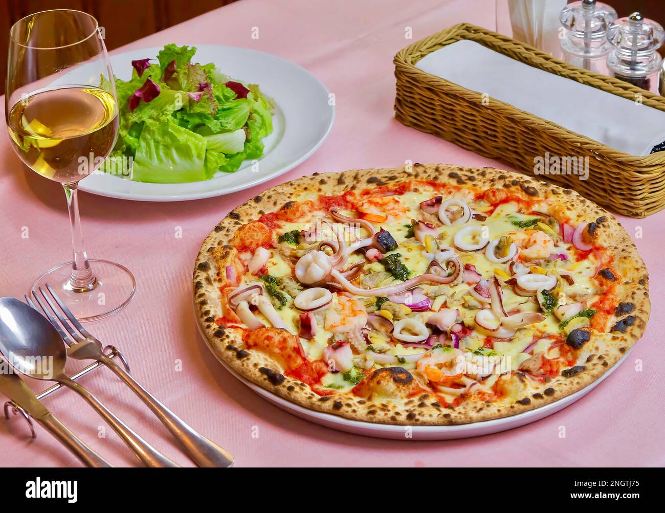 Pizza with seafood (Pizza ai frutti di mare) served with fresh lettuce and a glass of white wine. Isolated on a pink background. Close-up. Stock Photo