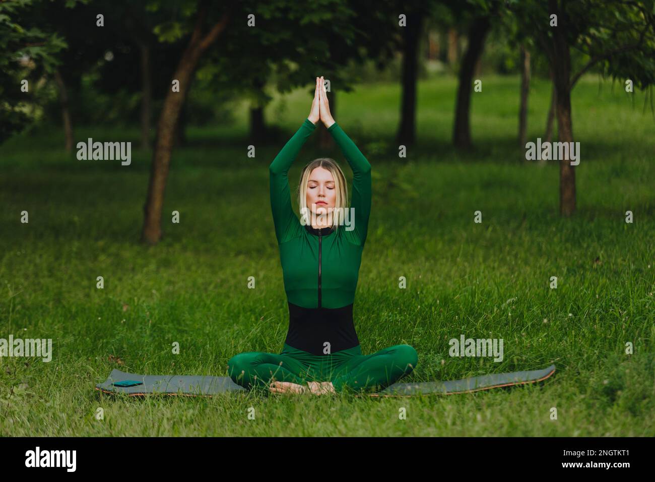 Yoga in the park with sunlight. A young woman in the lotus position sits on the green grass. The concept of calmness and meditation. Stock Photo