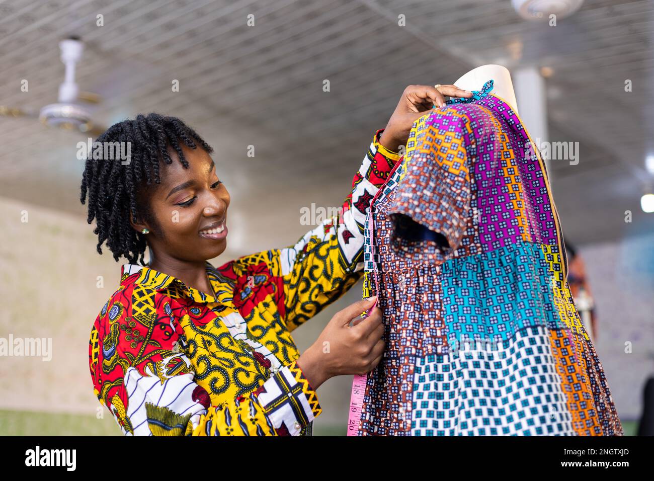 Smiling Ghanaian Dressmaker with locs hair measures a part of her colorful African pattern dress in her studio Stock Photo