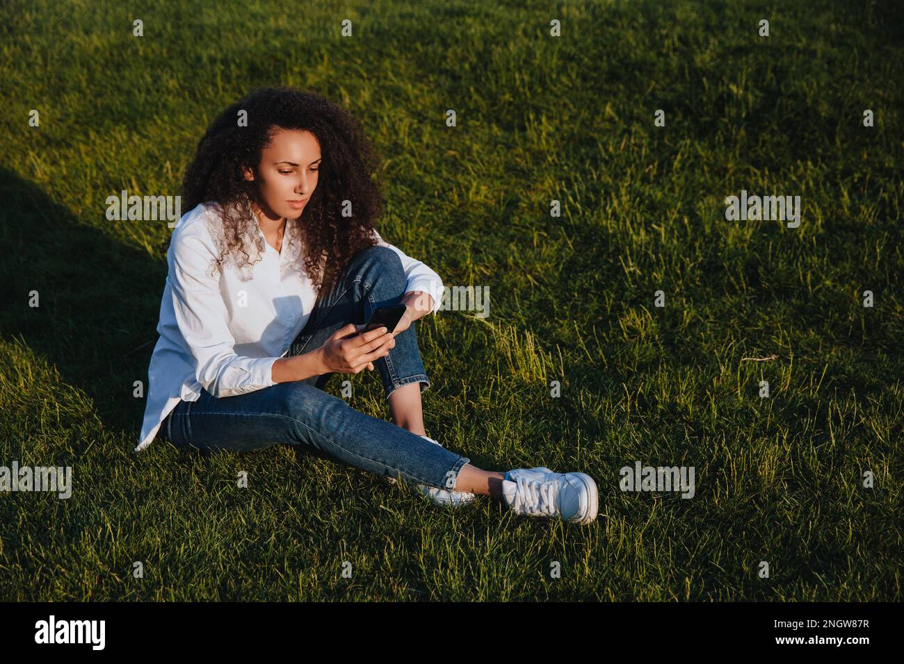 A young girl sits on the green grass in the park and uses social networks using a mobile phone. Stock Photo
