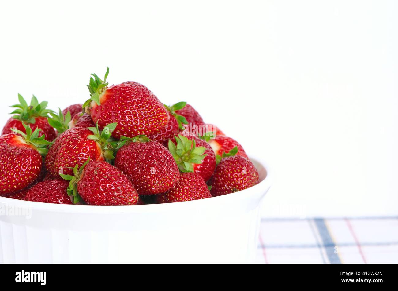 Fresh, whole ever-bearing strawberries (Fragaria x ananassa) in a white bowl on a blue plaid tablecloth with white background - copy space. Stock Photo