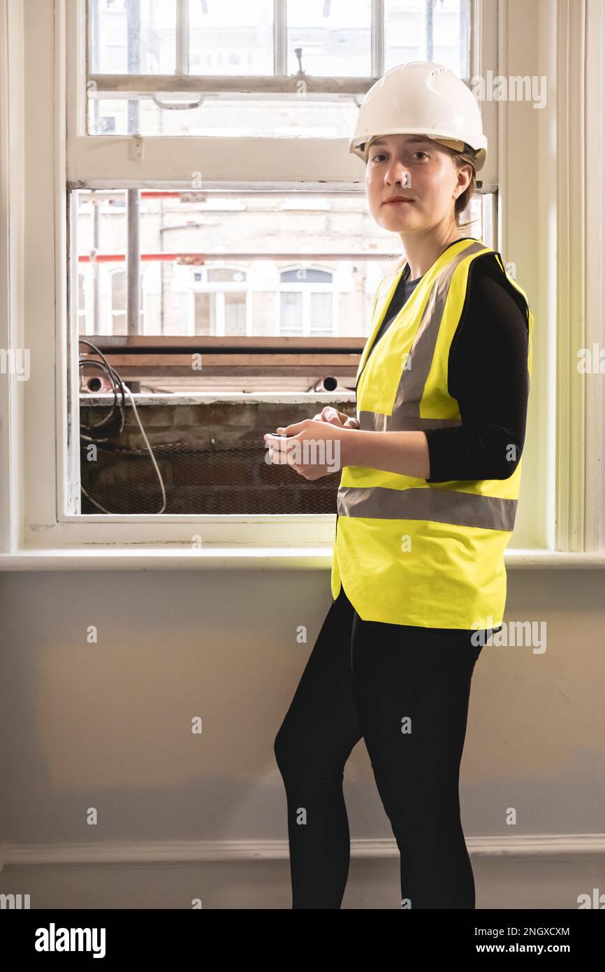 Vertical photo of a chartered civil engineer woman using a calculator in front of a window in an indoor construction site, wears hard hat, yellow ppe Stock Photo